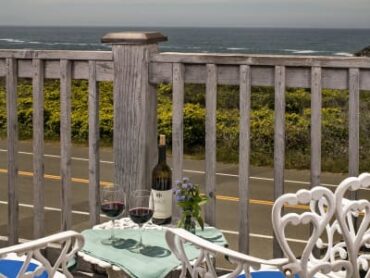 balcony overlooking road and water with white chairs, bottle and glasses of wine